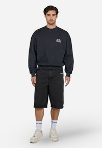 Sort sweatshirt med "PEGADOR" logo, parret med knælange sorte denimshorts. Fodtøjet er cremede sneakers, suppleret med hvide sokker.