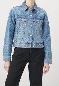 Chaqueta vaquera con patrón de logotipos en azul claro por todo el tejido, botones plateados, bolsillos con solapa en el pecho, combinada con pantalones negros en una persona de pie.