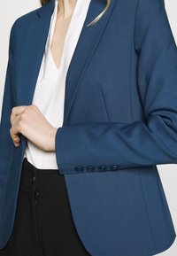 Blazer entallado azul con una textura suave, cierre de un solo botón y solapas con muesca sutil, usado sobre una blusa blanca y pantalones negros.