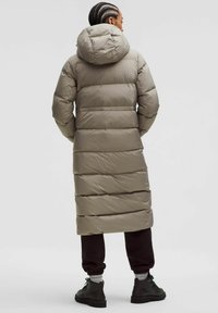 Dugi puffer kaput u svijetlo taupe boji s horizontalnim šavovima, velikim kapuljačom, bočnim džepovima i strukiranim strukom. Nositi preko tamnih hlača i čizama.