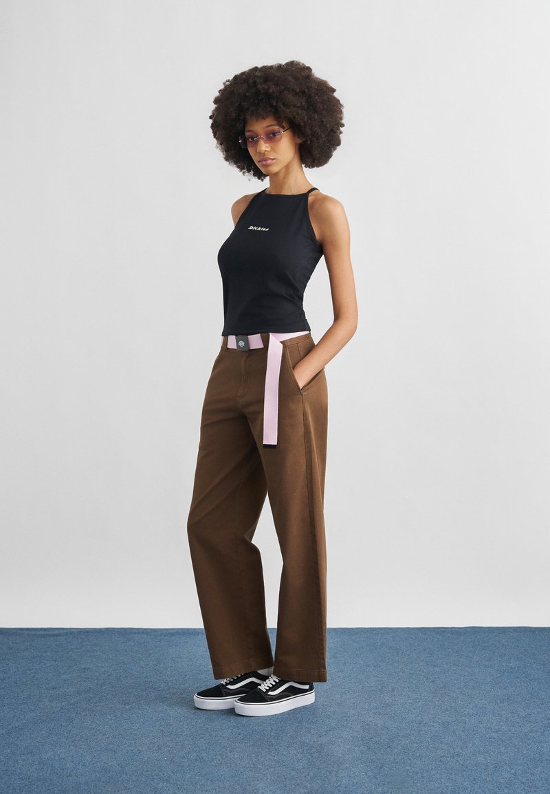 Woman with afro hairstyle wearing tinted glasses, black sleeveless top, brown wide-leg pants with pink belt, and black sneakers on blue floor.
