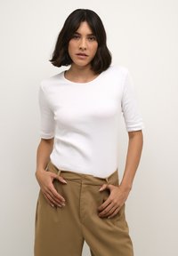 White fitted t-shirt with a round neckline and elbow-length sleeves, paired with high-waisted beige trousers. Smooth fabric texture.