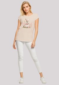 Beige short-sleeve T-shirt with a rabbit graphic surrounded by pink flowers, paired with slim white pants and white sneakers.