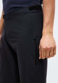 Άνδρας που φοράει μαύρο παντελόνι με φερμουάρ στην πλευρική τσέπη και ρυθμιζόμενη μέση, στέκεται με το χέρι χαλαρό στο πλάι.