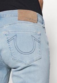 Vaqueros azul claro con un acabado desgastado. Presentan un parche de cuero marrón en la cinturilla con "TRUE RELIGION" grabado. Diseño icónico de bolsillo trasero con doble costura.