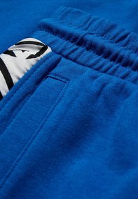 Siniset urheiluhousut, joissa on teksturoitu kangas, joustava vyötärö ja kontrastivärinen valko-musta logo-patch takataskussa.