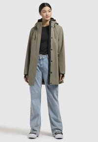 Impermeabile khaki con cappuccio, dotato di zip frontale e bottoni a pressione. Abbinato a jeans azzurri larghi e scarpe nere.