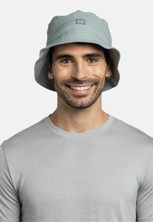 Smiling man with short beard wearing light gray bucket hat with logo, matching light gray crew neck t-shirt, facing forward on white background.