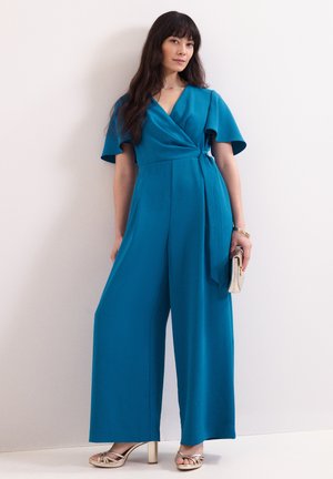 Femme aux longs cheveux foncés portant une combinaison bleue à jambes larges, tenant une pochette beige, debout contre un mur blanc.