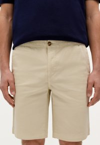 Beige shorts med elastisk talje og en brun knapdetalje. Stoffet er glat, letvægts og har sidelommer.