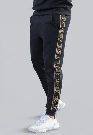 Czarne zwężane spodnie dresowe z złotymi paskami z logo "SIKSILK" po bokach, noszone z białymi teksturowanymi trampkami.