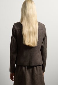 Donna con lunghi capelli biondi, che indossa una giacca e una gonna marrone scuro, in piedi contro un muro chiaro e uniforme, voltata di spalle alla telecamera.