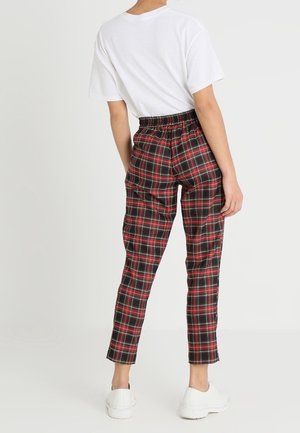 Osoba nosící bílé tričko, červené a černo kostkované kalhoty s elastickým pasem a bílé tenisky, stojící před světlým jednobarevným pozadím.