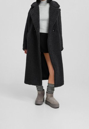 Personne portant un long manteau noir texturé, un pull à col roulé gris clair, une jupe noire, des chaussettes grises et des bottines grises sur un fond uni.
