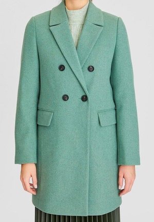 Kobieta w dwurzędowym, miętowozielonym wełnianym płaszczu z klapami z wycięciami i kieszeniami z patkami, w połączeniu z jasnoszarym golfem.