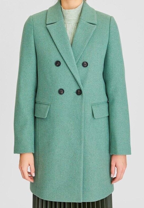 Femme portant un manteau en laine vert menthe à double boutonnage avec des revers crantés et des poches à rabat, associé à un pull à col roulé gris clair.