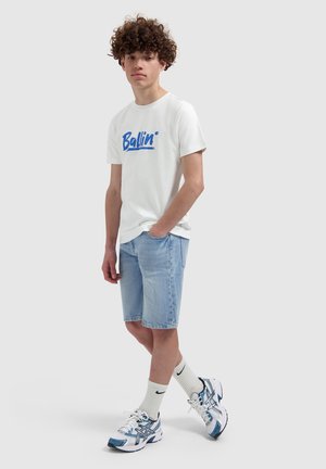 Jonge man met krullend haar die een wit "Ballin" T-shirt draagt, lichte denimshorts, witte Nike-sokken en wit-blauwe sneakers, staand met één hand in zijn zak.