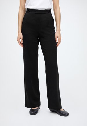 Femme portant un pantalon noir taille haute à jambes larges avec des détails à boutons et des chaussures plates noires sur fond blanc.