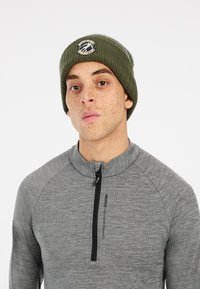 Groene gebreide beanie met een logo-opdruk, gedragen met een grijs lange mouw zip-up shirt met een gestructureerd patroon en een ritskraag.