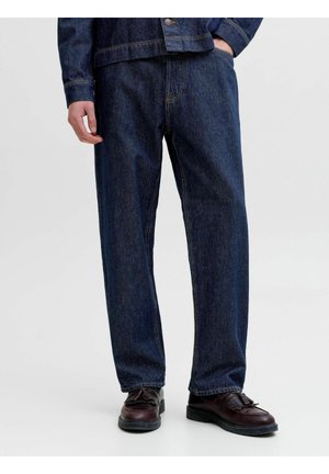 Person trägt dunkelblaue Jeans und passende Jeansjacke, mit einer Hand in der Tasche und braunen Loafers mit Quasten.