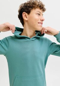 Ung person med krøllet hår, der justerer hætten på en teal sweatshirt, smilende og kigger til højre mod en neutral baggrund.