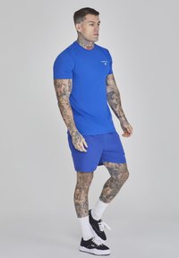 Mėlyna marškinėliai ir mėlyni šortai, pagaminti iš minkštos medžiagos. Ant rankų ir kojų yra tatuiruočių, juodi sportbačiai ir balti kojinės. Neutrali fono spalva.