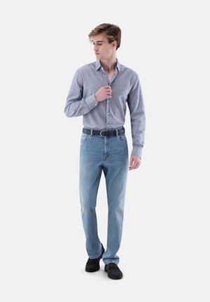Junger Mann trägt ein blau gestreiftes Hemd mit Knöpfen, hellblaue Jeans, einen schwarzen Gürtel und schwarze Schuhe, richtet seinen Hemdkragen und schaut nach links.