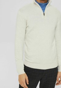 Helles beige Zip-Knit-Pullover aus weichem Strickstoff, mit langen Ärmeln und geripptem Saum. Über einem blauen Rollkragenpullover getragen.