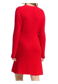 Femme portant une robe rouge en maille côtelée à manches longues, vue de dos, avec un ourlet légèrement évasé au-dessus des genoux.