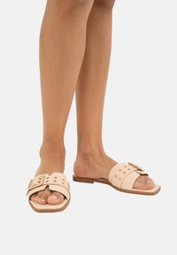 Sandali scivolosi beige con una fascia larga, caratterizzati da dettagli metallici in tonalità oro e borchie. La suola piatta e il design open-toe mettono in risalto le dita dei piedi.