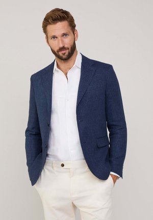 Veste à rayures navy avec revers crantés, fermeture à un bouton et deux poches avant, portée sur une chemise blanche et un pantalon clair.