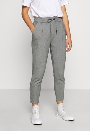 Person iført grå joggingbukser med snøre, hvide høje sneakers og en hvid T-shirt stoppet ned i bukserne, stående mod en ensfarvet baggrund.