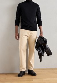 Pull à col roulé noir, pantalon couleur crème, et veste en cuir noire tenue à la main. Des bottines noires complètent la tenue sur un sol en bois.