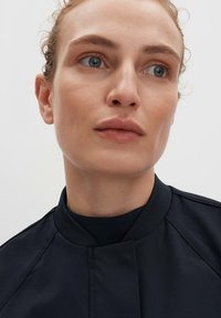 Close-up of a person wearing a dark navy jacket featuring a high collar and smooth fabric. Light skin tone and neutral makeup.