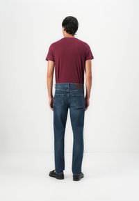 Hombre vistiendo una camiseta color burdeos, jeans azules y zapatos negros de pie frente a una pared blanca con las manos relajadas a los lados.