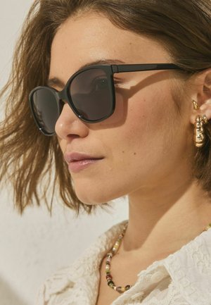 Junge Frau mit kurzen braunen Haaren, die schwarze Sonnenbrillen, goldene Ohrringe, eine bunte Perlenkette und ein weißes, strukturiertes Oberteil trägt.