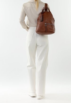 Person wearing beige shirt and white pants, carrying large brown leather backpack over right shoulder, standing against white background.