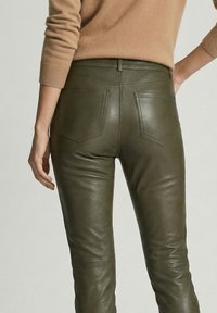 Persoon die olijfgroene leren broek en een camelgekleurde trui met lange mouwen draagt, gezien vanaf de achterkant ter hoogte van de taille.