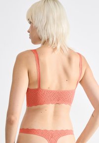 Koronkowy bralette w kolorze koralowym z geometrycznym wzorem, falowanymi krawędziami i cienkimi ramiączkami, zestawiony z pasującymi koronkowymi stringami. Gładka faktura.