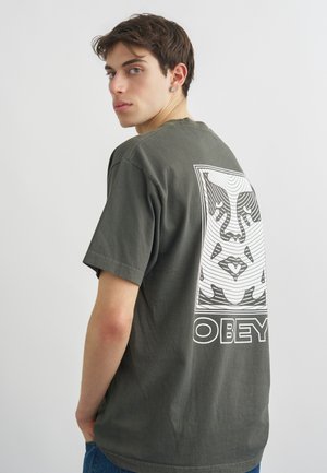 Ung mand med kort brunt hår iført en mørkegrå OBEY t-shirt med en stor stiliseret ansigtsgrafik på ryggen, der kigger over skulderen.