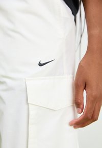 Bijele cargo hlače s teksturiranim završetkom, s velikom bočnom džepom i malim crnim Nike logotipom na prednjoj strani.