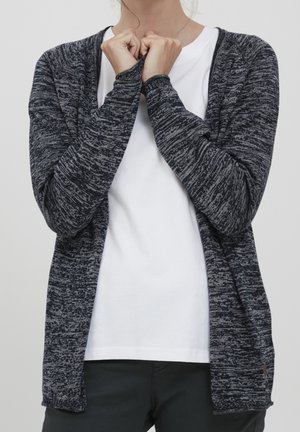 Woman wearing a dark gray marled cardigan over a white t-shirt, holding the cardigan collar with both hands, standing against a plain background.