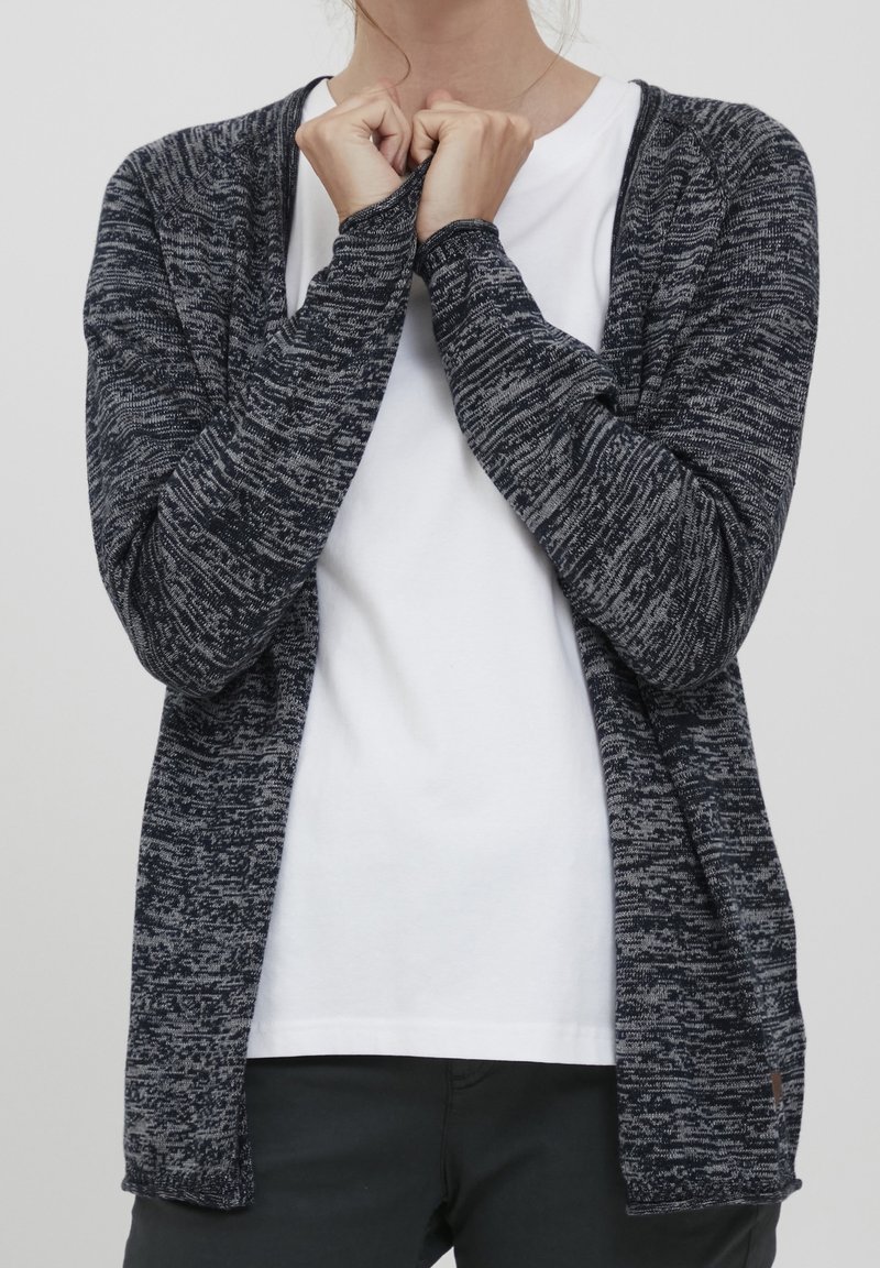 Femme portant un cardigan gris foncé chiné par-dessus un t-shirt blanc, tenant le col du cardigan avec les deux mains, debout devant un fond uni.