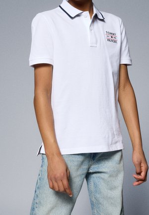 Persoon draagt een witte Tommy Hilfiger polo met zwarte bies op de kraag en lichtblauwe spijkerbroek, staand tegen een effen achtergrond.