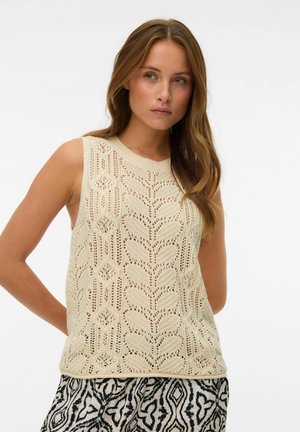 Mlajša ženska z dolgimi lasmi, oblečena v brezrokavnik iz bež pletenine in črno-belo krilo s potiskom, stoji pred enobarvnim ozadjem.