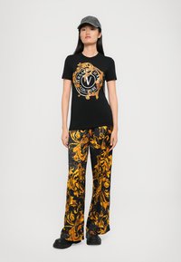 Woman wearing black cap, black Versace Jeans Couture t-shirt with gold ornate design, matching black and gold patterned pants, and black shoes.