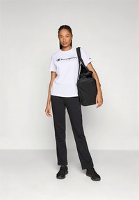 White short-sleeve t-shirt with "Champion" logo, black pants, and black shoes. Carrying a black bag, standing on a light gray background.