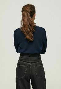 Frau mit langen braunen Haaren im Pferdeschwanz, die einen marineblauen Pullover und hoch taillierte schwarze Jeans trägt, stehend vor einem schlichten hellen Hintergrund.