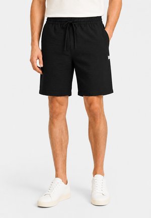 Man wearing black drawstring shorts, white T-shirt, and white sneakers, standing with one hand in pocket against a plain background.