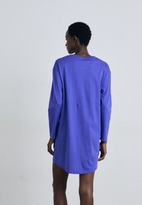 Vestido camiseiro de manga longa roxo, feito de algodão suave, com um corte descontraído e colarinho redondo, terminando a meio do thigh, com um design simples.
